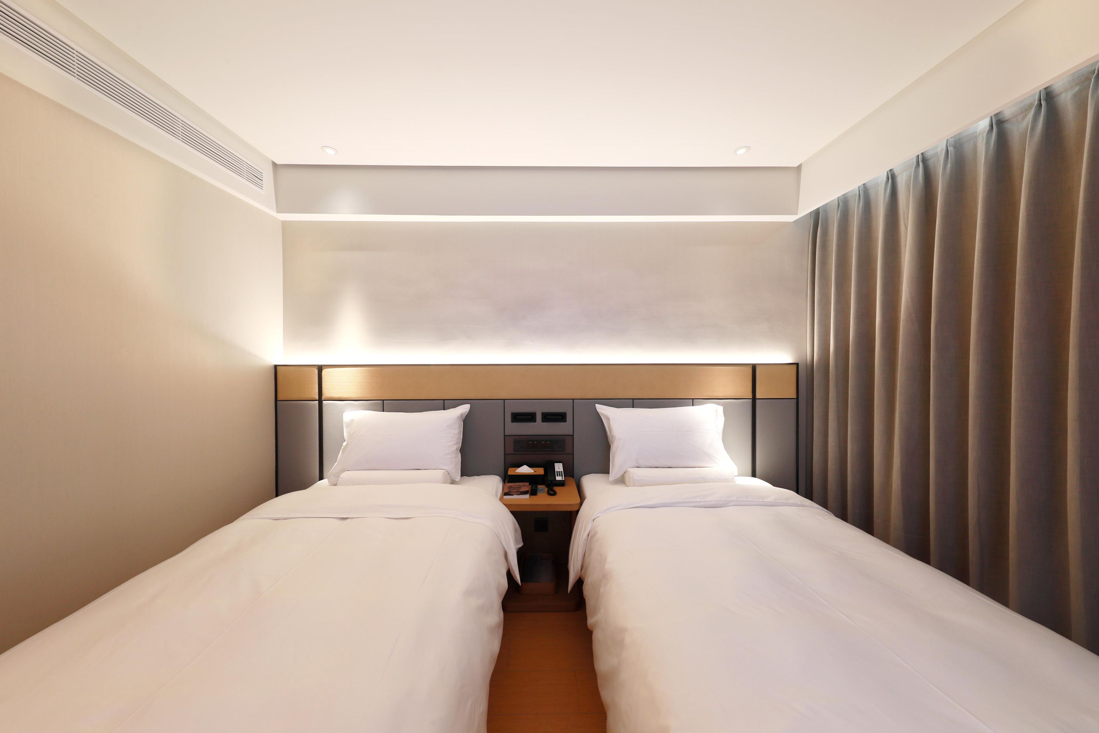 A hotel room with two single beds, a bedside table with a lamp, and a large window with curtains.