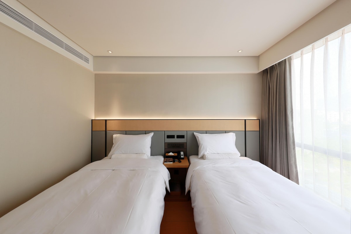 Habitación de hotel con dos camas individuales, una mesita de noche con lámpara, un pequeño escritorio con teléfono y una ventana con cortinas.