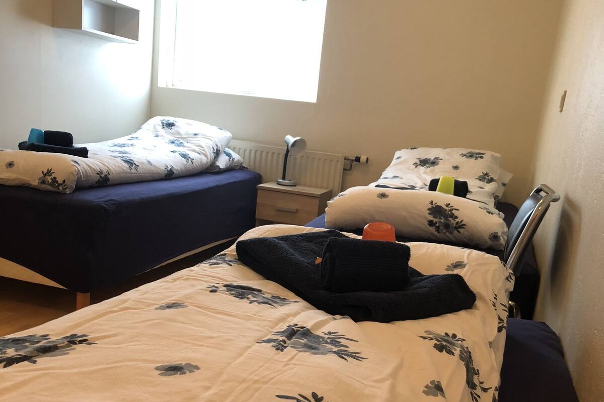 Ein Schlafzimmer mit zwei Betten, eines mit blauer Bettdecke und das andere mit beiger Bettdecke. Ein Nachttisch mit Lampe und ein kleiner Tisch mit einer Flasche darauf.