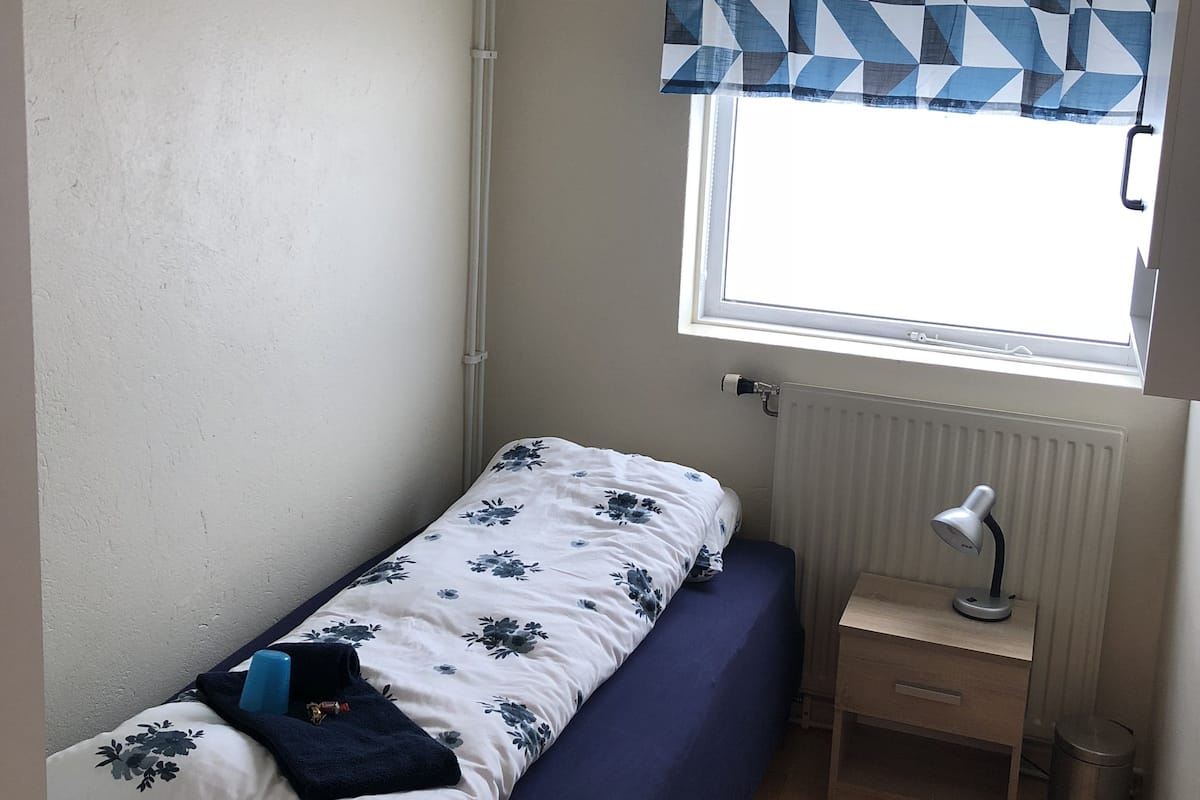 Ein kleines Zimmer mit einem Einzelbett, einem Fenster, einem Nachttisch mit Lampe und einer Heizung.