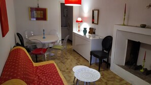 Een woonkamer met een bank, stoelen, een eettafel, een klein witte tafel, een open haard en een keuken met een aanrecht en kastjes.