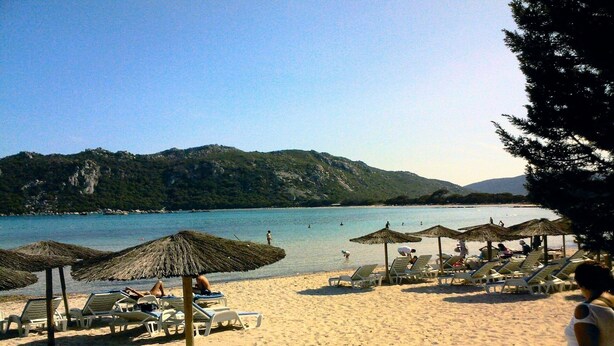 Een strand met rieten parasols en ligbedden, een rustige zee en een bergachtige achtergrond.