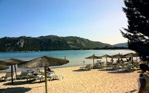 Ein Strand mit Reetdachschirmen und Liegestühlen, ruhigem Meer und einem bergigen Hintergrund.