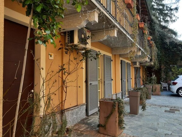 A row of terracotta buildings with closed shutters, a balcony with plants, and a parked car. - B&B Corte delle Rose (Voghera)