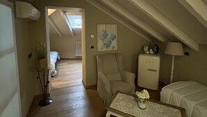Un dormitorio acogedor en el desván, con techo inclinado, piso de madera, cama, sillón, una mesita con un florero y un radiador.