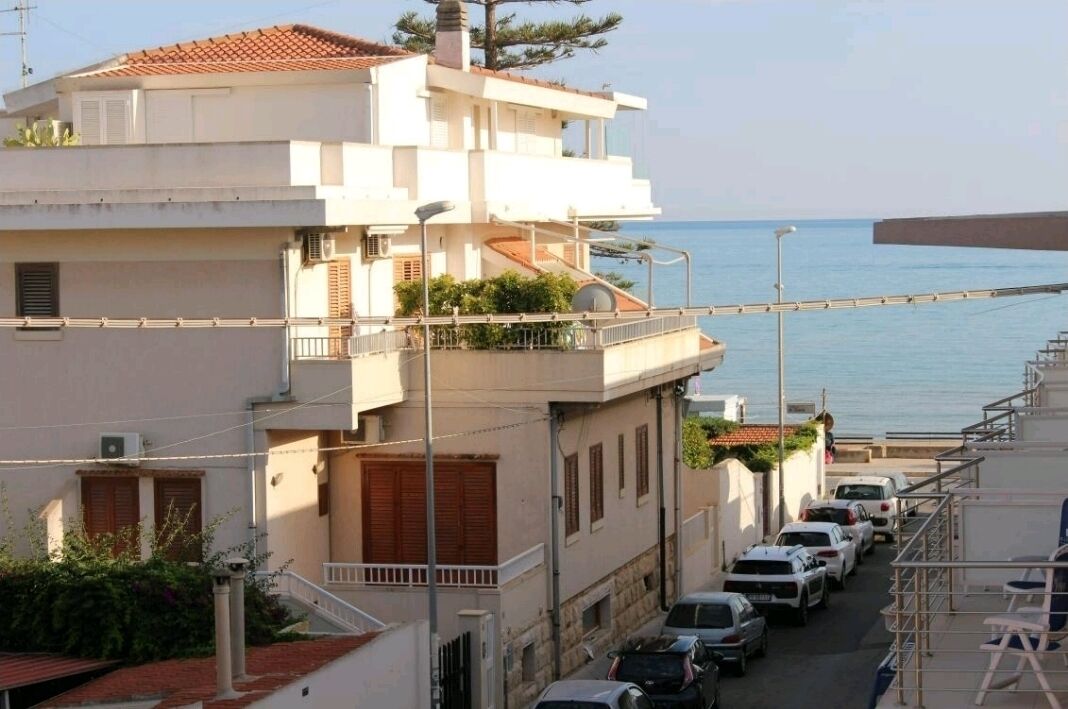 A coastal residential building with a balcony overlooking the sea, parked cars along the street, and a clear view of the ocean.