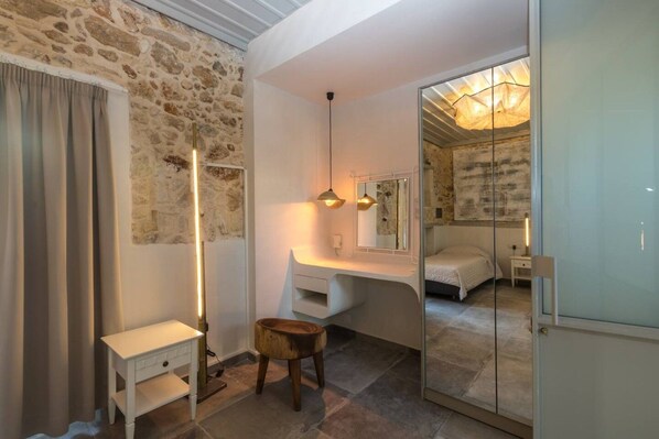 A bedroom with a stone wall, a bedside table, a mirror, a bed, and a glass door leading to another room.