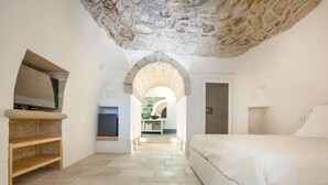 A bedroom with a stone ceiling, a TV on a wooden shelf, and a view to a kitchen and bathroom through an archway. - Luxury Villa Le Muse between Santa Maria al Bagno and Nardò with swimming pool and Wifi (Santa Maria al Bagno)