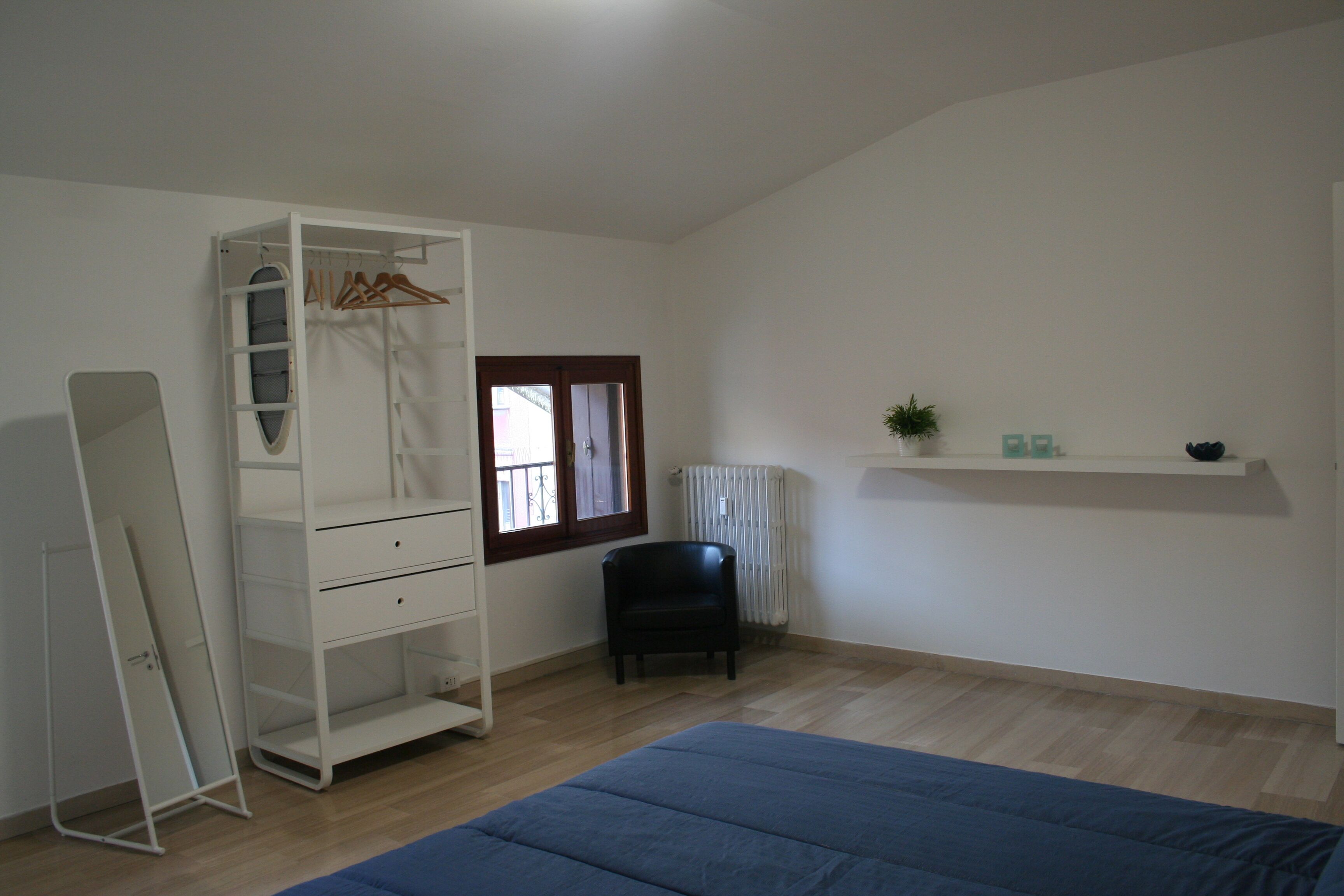 Een kamer met een bed, een kledingkast, een stoel, een radiator en een plank met spullen.