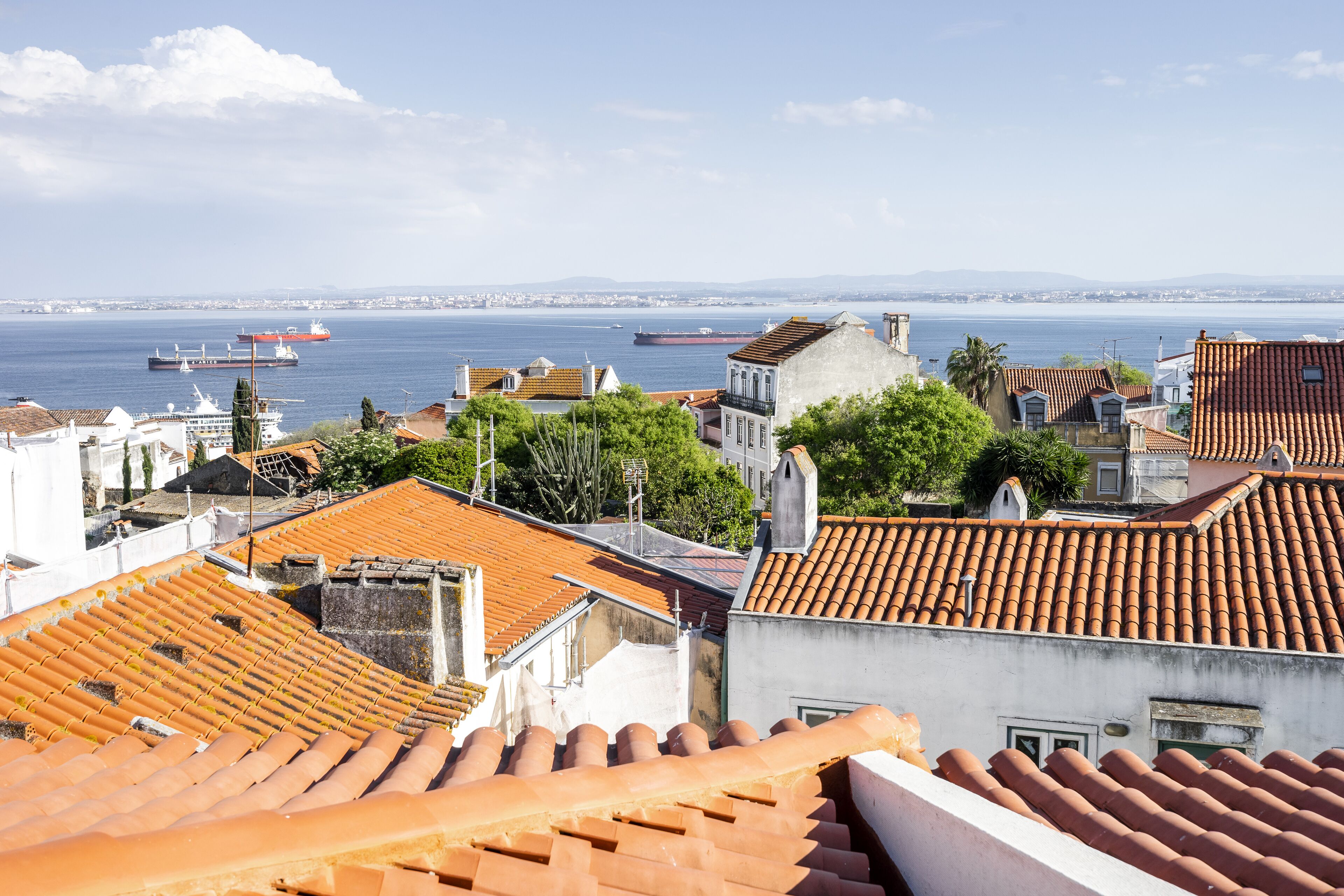 Un paisaje urbano costero con edificios de techo de tejas color terracota, un cielo azul despejado y un mar tranquilo con barcos.