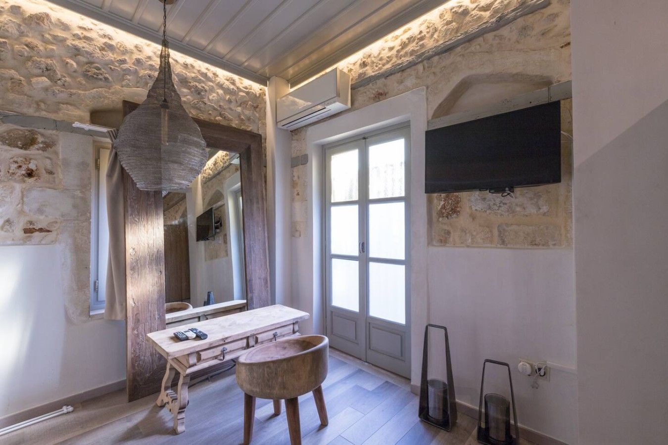 A bathroom with a stone wall, wooden floor, a wooden vanity, a hanging light fixture, and a large window.