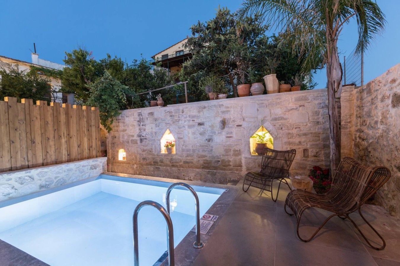 A swimming pool with a stone wall, two wicker chairs, and a wooden fence.