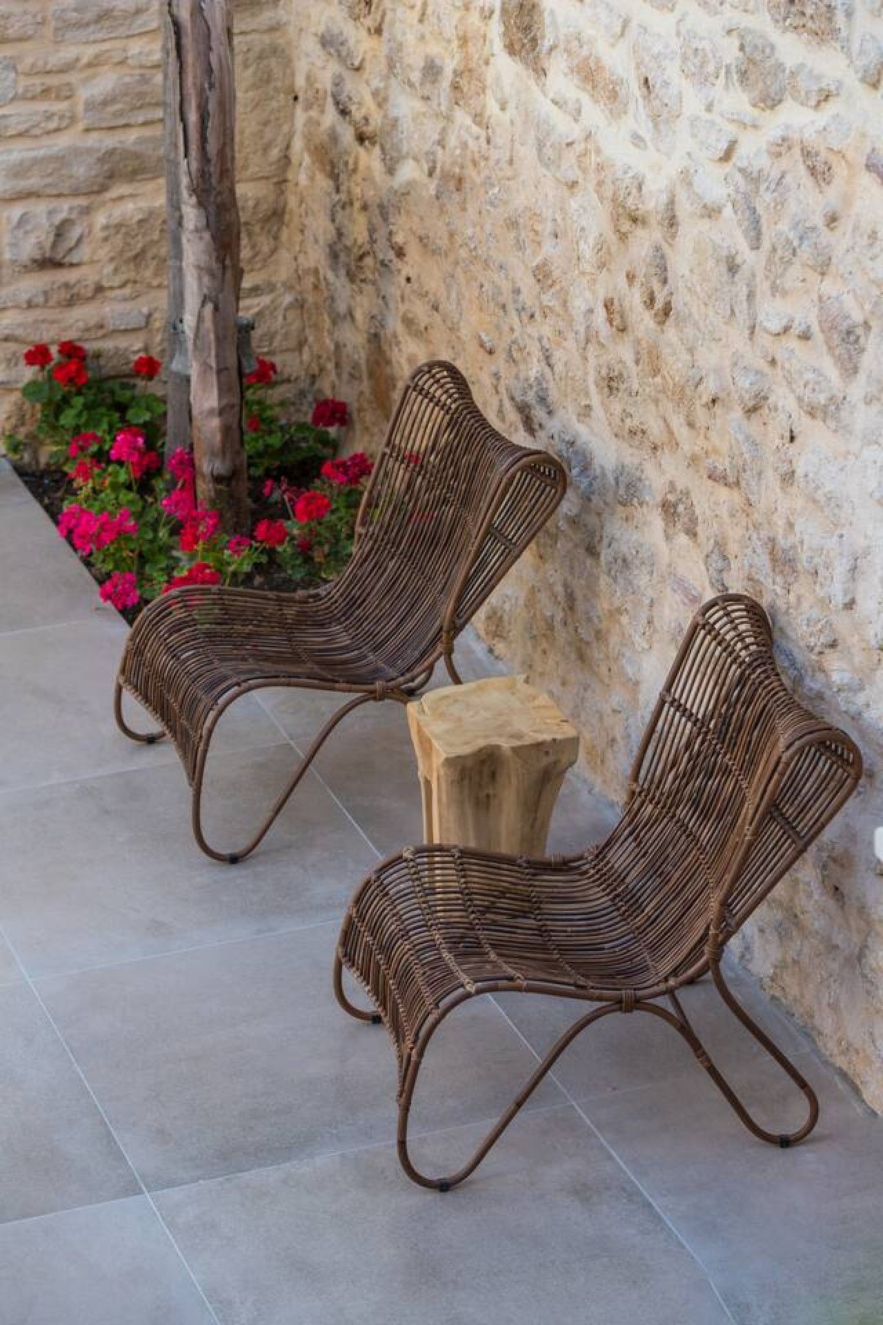 Two wicker chairs with metal frames, positioned on a tiled patio next to a stone wall with a wooden stump and red flowers.