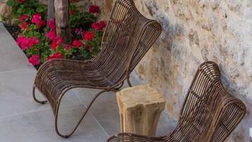 Two wicker chairs with metal frames, positioned on a tiled patio next to a stone wall with a wooden stump and red flowers.