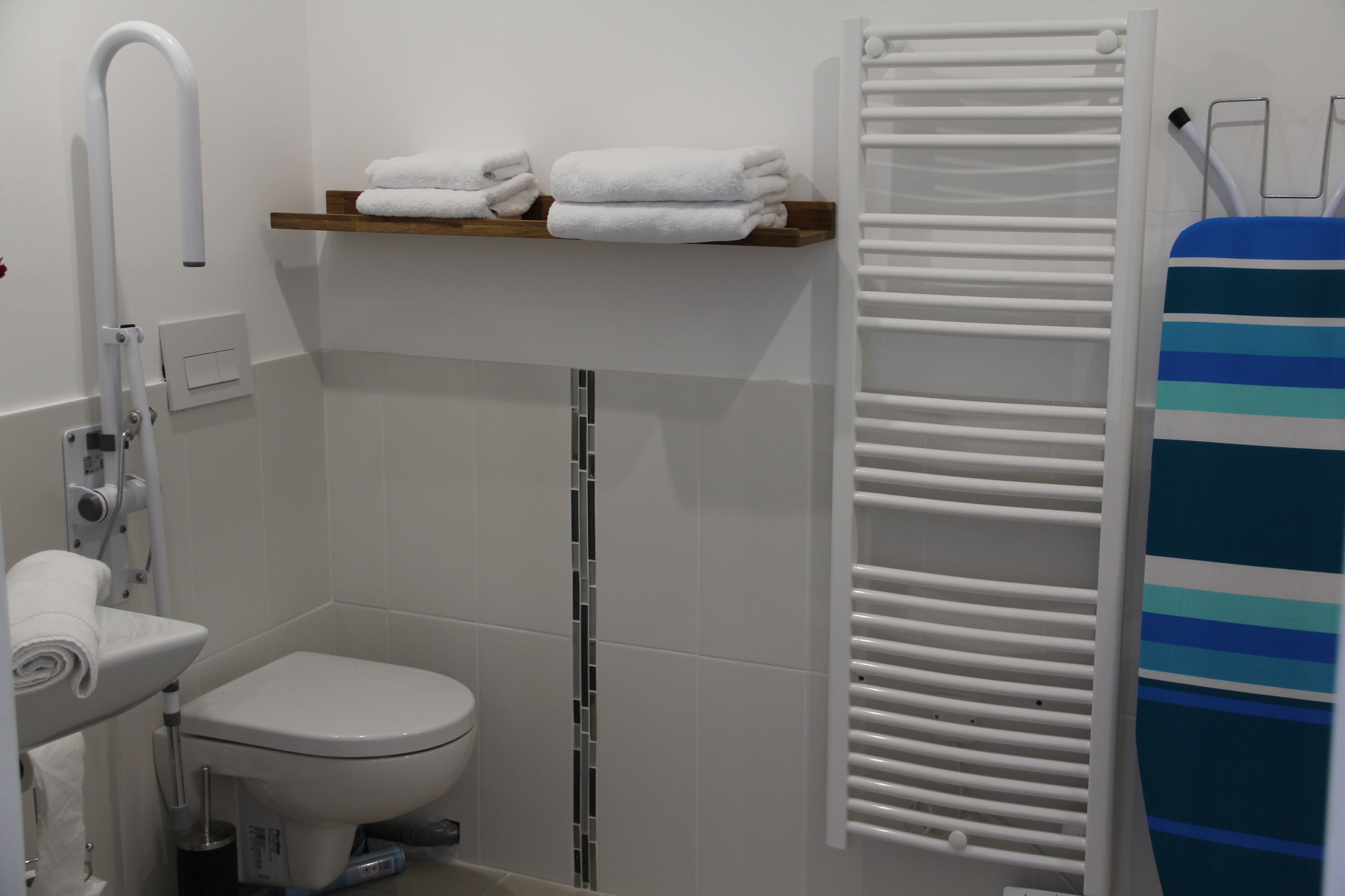 Ein Badezimmer mit Toilette, Ablagefläche mit gefalteten Handtüchern, weißem Handtuchhalter und Duschbereich mit blauem Handtuch.