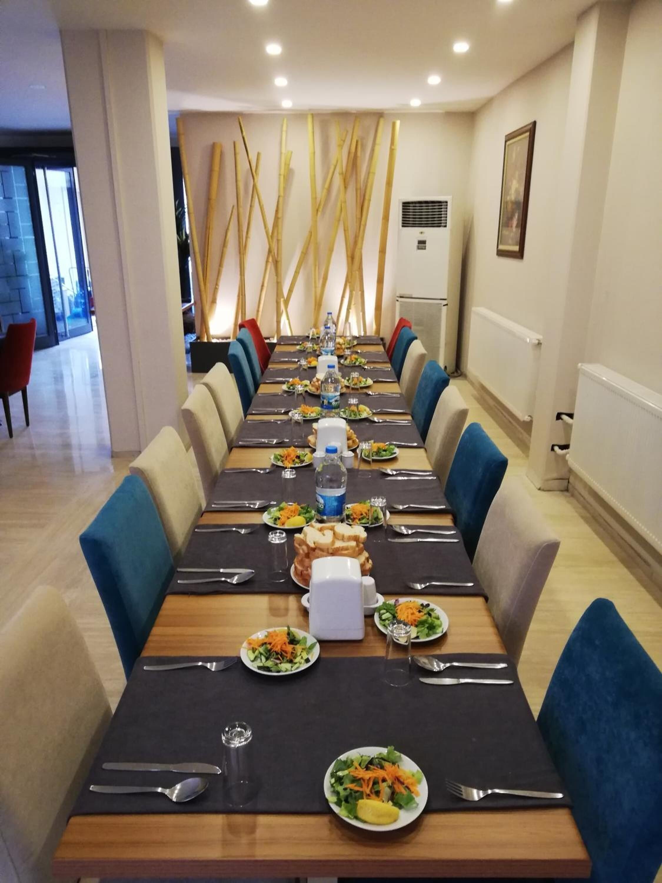 A long dining table set with plates, cutlery, and water bottles, flanked by blue upholstered chairs in a modern interior with a bamboo wall feature.