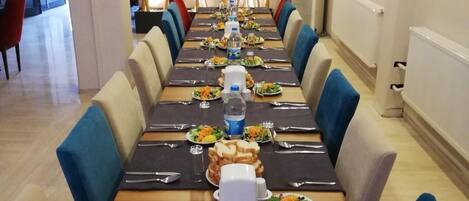 A long dining table set with plates, cutlery, and water bottles, flanked by blue upholstered chairs in a modern interior with a bamboo wall feature.