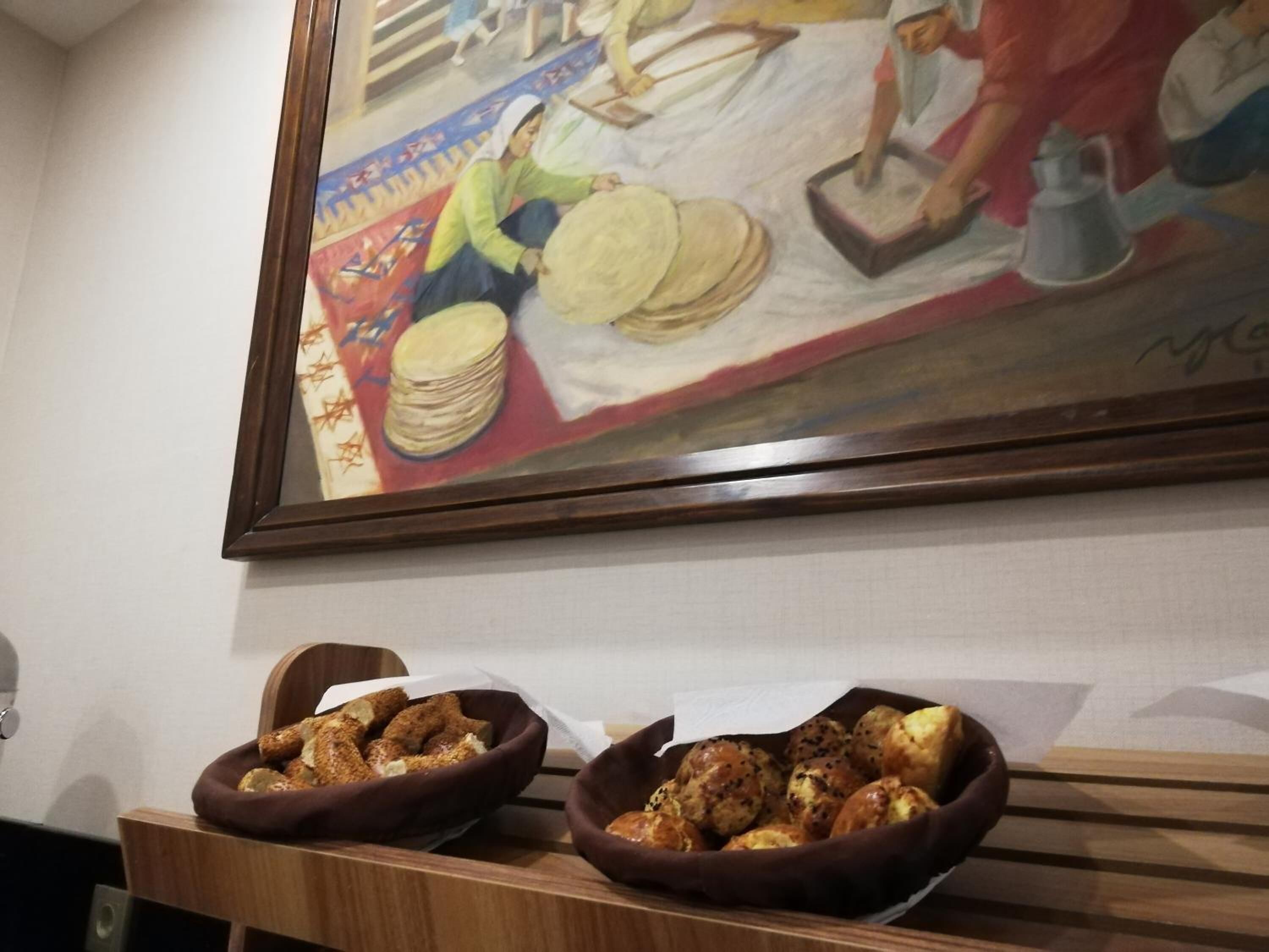 A room with a painting of women working on a textile, two bowls of bread on a wooden table.