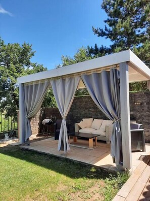 Een overdekt terras met een bank en een tafel, omringd door een stenen muur en groen.