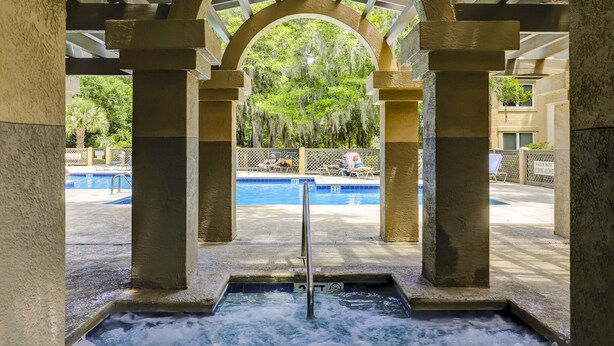 Área de piscina com hidromassagem sob uma arcada coberta, rodeada por espreguiçadeiras e vista para um jardim exuberante.