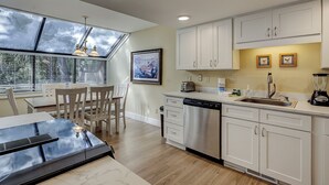 A modern kitchen with white cabinets, a dishwasher, and a dining area with a table and chairs. - Sunset Rentals | Beachwalk 110 (Hilton Head)