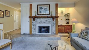 A living room with a stone fireplace, wooden mantel, and a framed picture above it. There is a sofa and a chair in the room. - Sunset Rentals | Beachwalk 110 (Hilton Head)