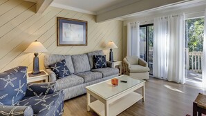 A living room with a sofa, two side tables with lamps, a glass coffee table, and a view of the outdoors through a sliding glass door. - Sunset Rentals | Beachwalk 110 (Hilton Head)