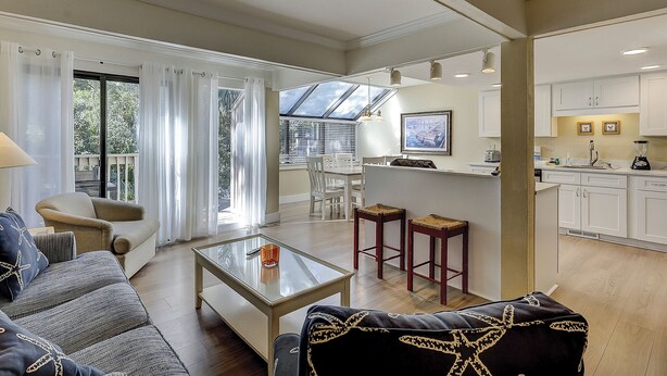 A spacious living area with a sofa, a glass coffee table, and a dining table with chairs. There is a kitchen area with white cabinets and a window overlooking the garden. - Sunset Rentals | Beachwalk 110 (Hilton Head)