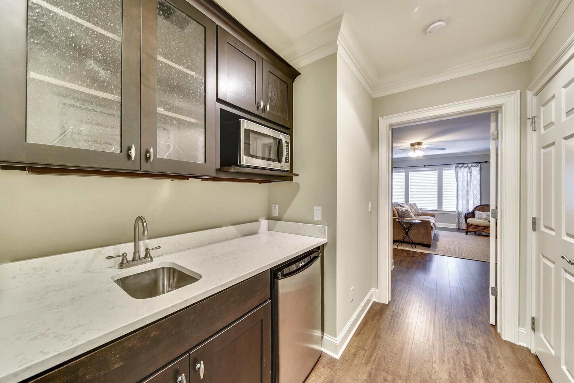 A kitchen with dark cabinets, a stainless steel sink, and a dishwasher. There is a hallway leading to a living area with a couch and a ceiling fan.