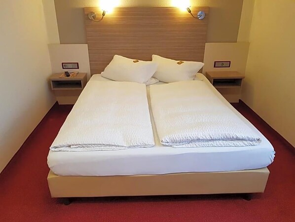 A double bed with white linens, two bedside tables, and a wooden headboard with wall-mounted lights.