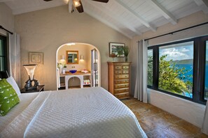 A bedroom with a bed, a vanity with a mirror, a dresser, and a large window with a view of the sea.