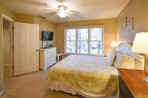 A bedroom with a bed, a television on a dresser, a window with a view of trees, and a ceiling fan.