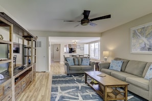 A living room with a sofa, coffee table, and a ceiling fan. There is a kitchen in the background with a dining area.