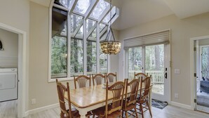 A dining room with a large wooden table and chairs, a chandelier, and large windows with a view of trees. - Sunset Rentals | Mooring Buoy 175 (Hilton Head Island)