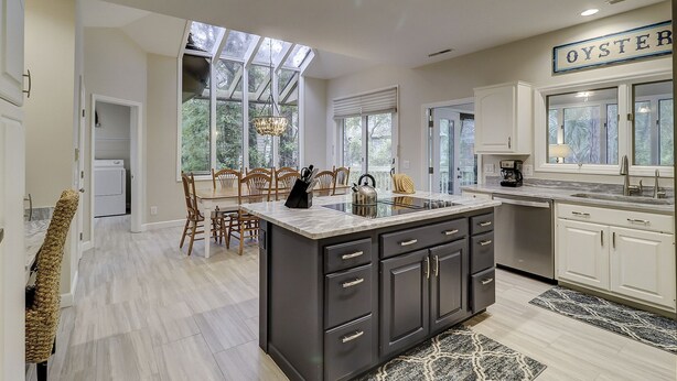 A modern kitchen with a central island, white cabinetry, and a view of the outdoors through large windows. - Sunset Rentals | Mooring Buoy 175 (Hilton Head Island)