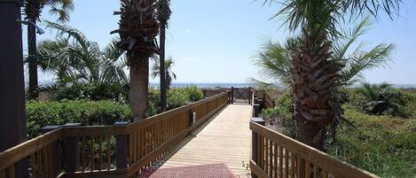 A wooden boardwalk with metal grates, surrounded by palm trees and greenery, leading to a beach.