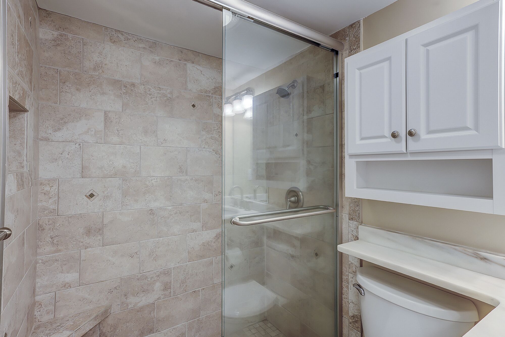 A bathroom with a glass-enclosed shower, beige tiles, a white toilet, and a white cabinet with drawers.