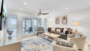 A spacious living room with a glass coffee table, a sofa, and a chair. There is a ceiling fan, a TV, and artwork on the wall.