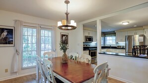 A dining area with a wooden table and white chairs, a chandelier, and a view into the kitchen with modern appliances. - Sunset Rentals | Evian 257 | Dog Friendly (Hilton Head Island)