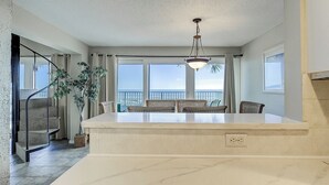 Une cuisine spacieuse avec un grand îlot central, un coin repas avec des chaises en osier, un escalier et une vue sur l’océan.