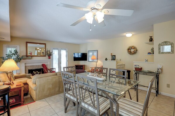 A living room with a fireplace, a television, and a dining area with a glass table and chairs. - Sunset Rentals | Evian 103 | Dog Friendly (Hilton Head)
