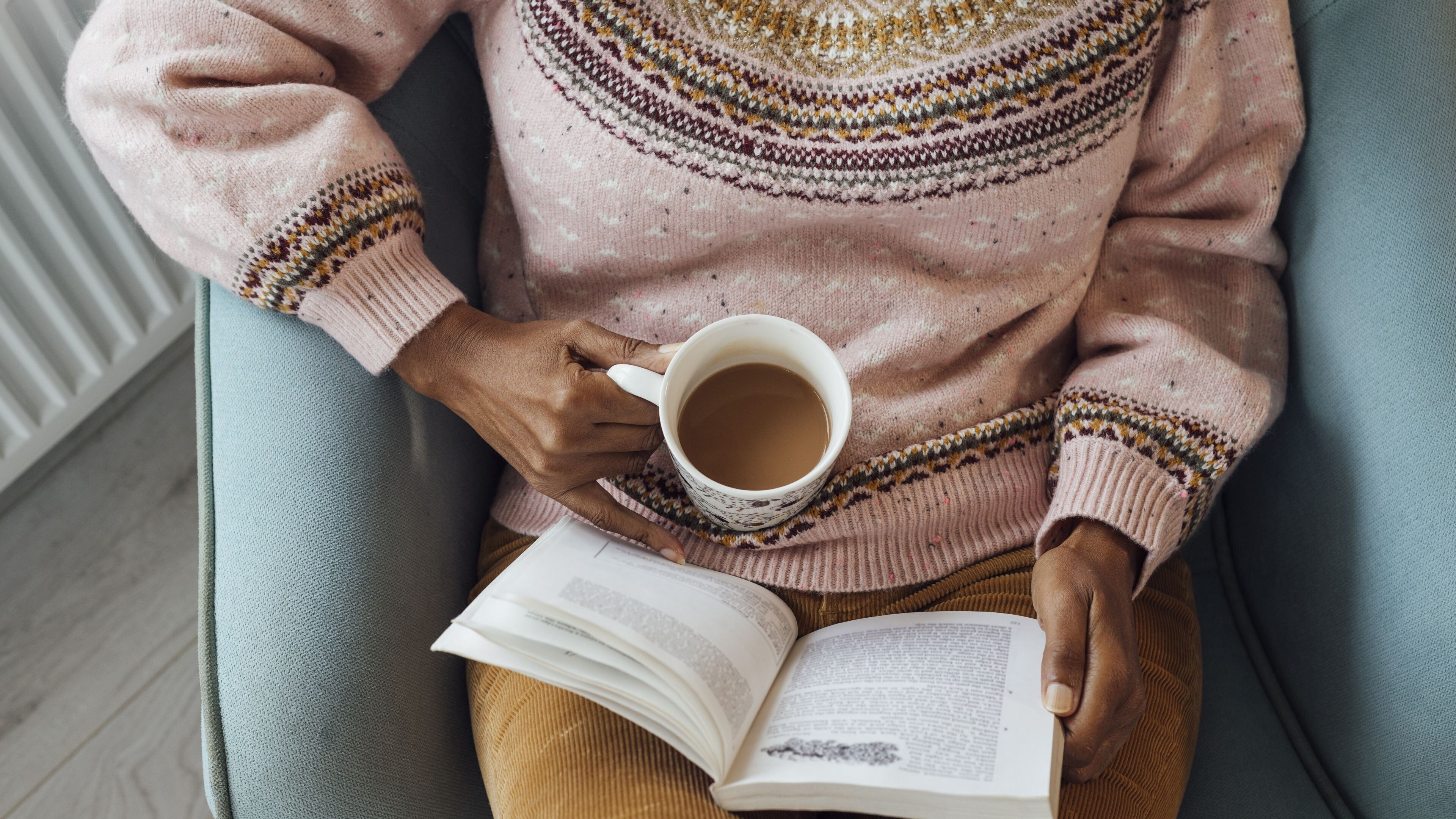 A person sitting on a chair, holding a book and a cup of coffee.