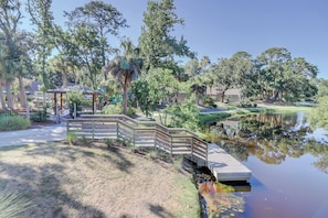 Stroll along the tranquil boardwalk over a serene body of water, surrounded by lush trees and manicured lawns.