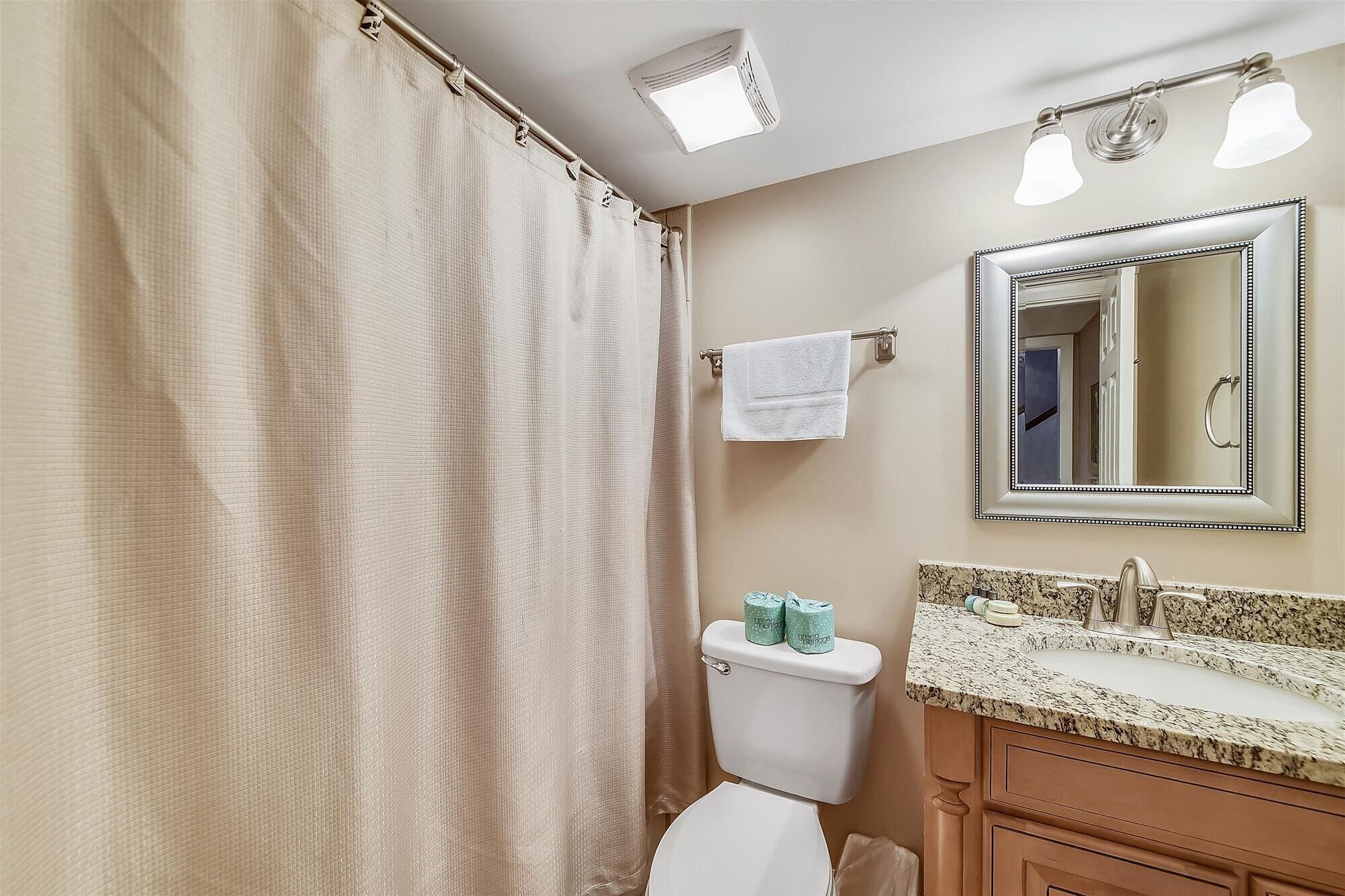 Une salle de bain équipée d’une douche avec rideau, de toilettes, d’un lavabo avec un plan de travail en granit et d’un miroir avec un cadre décoratif.