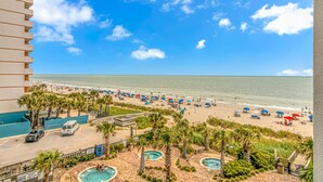 A beach view from a hotel balcony, including a swimming pool and palm trees. - Palace 1404 (Myrtle Beach)