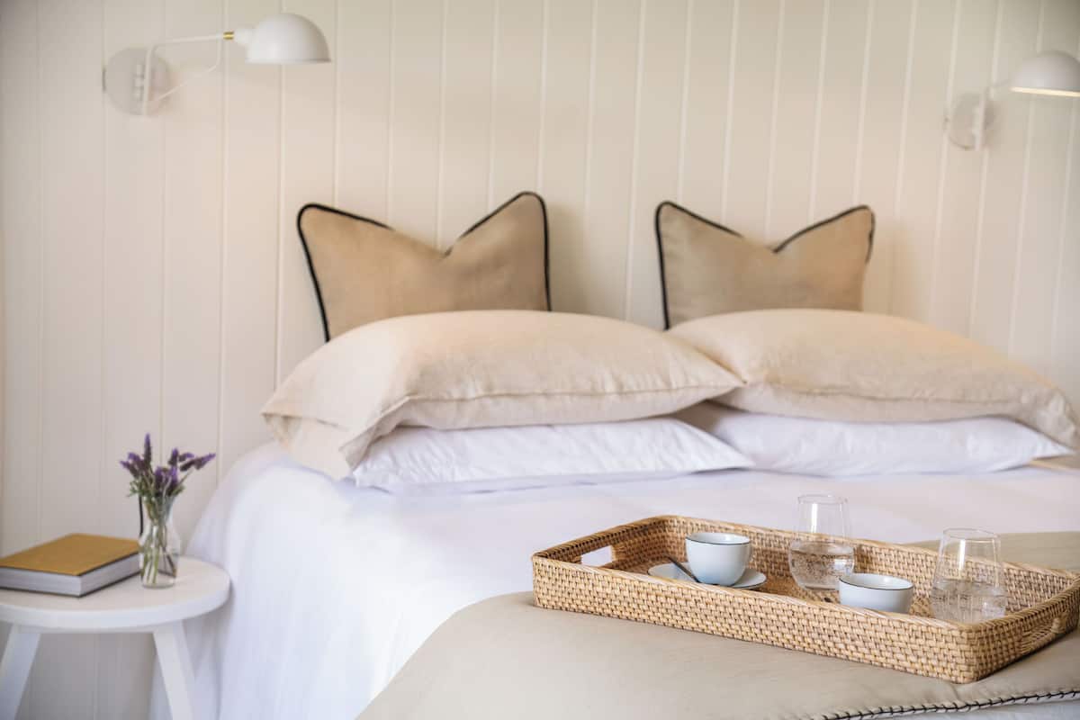 Cama bem arrumada com duas almofadas, mesa de cabeceira com um livro e um vaso de flores, e bandeja com copos e tigelas na cama.