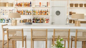 A bar area with white shelves stocked with bottles and glasses, wicker chairs, and a white table with a vase of greenery. - Posada Arenas de José Ignacio (José Ignacio)