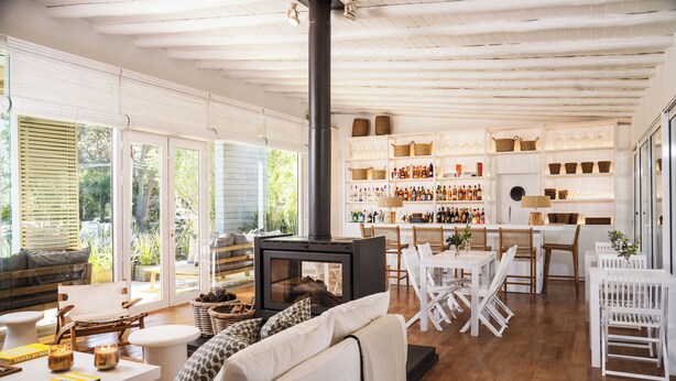 A spacious living area with a fireplace, wooden furniture, and a kitchen area with shelves of bottles. - Posada Arenas de José Ignacio (José Ignacio)