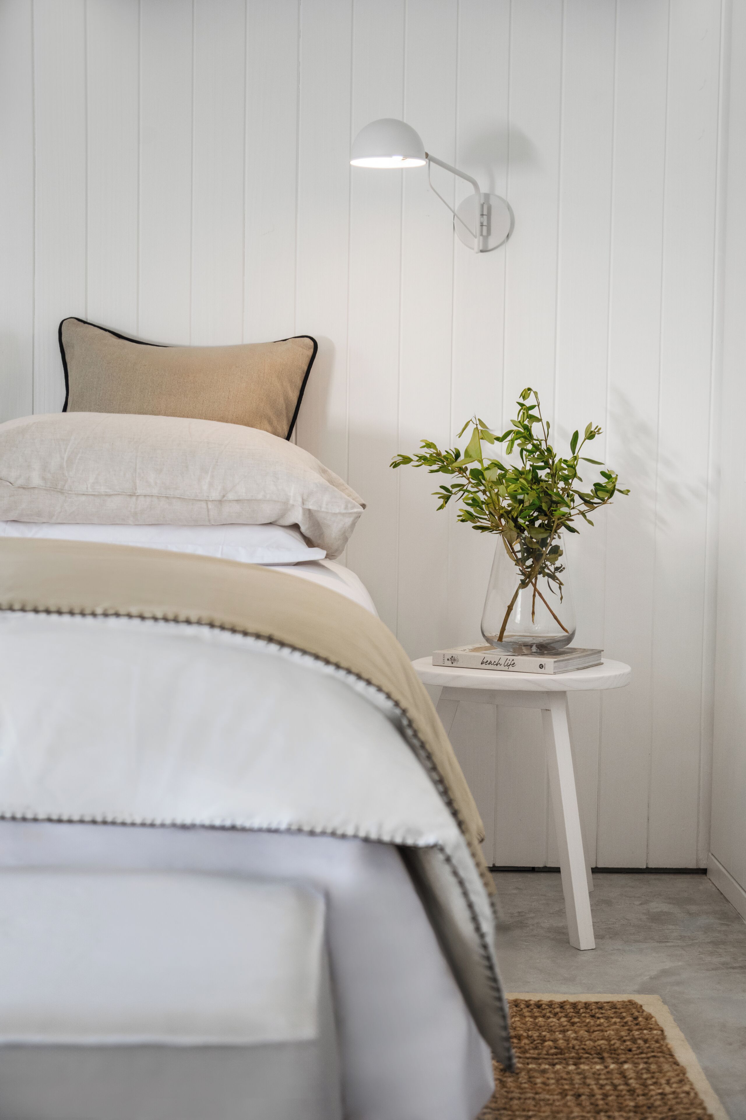 Una cama bien hecha con edredón beige y blanco, una mesita de noche pequeña en blanco con un florero de plantas verdes, y una lámpara fijada a la pared.