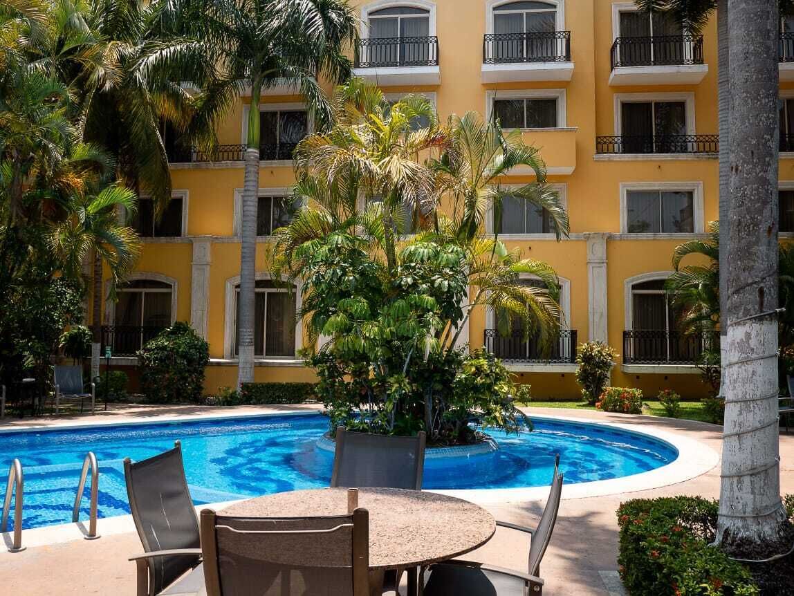 A hotel pool area with a circular pool, palm trees, and a yellow building with balconies.